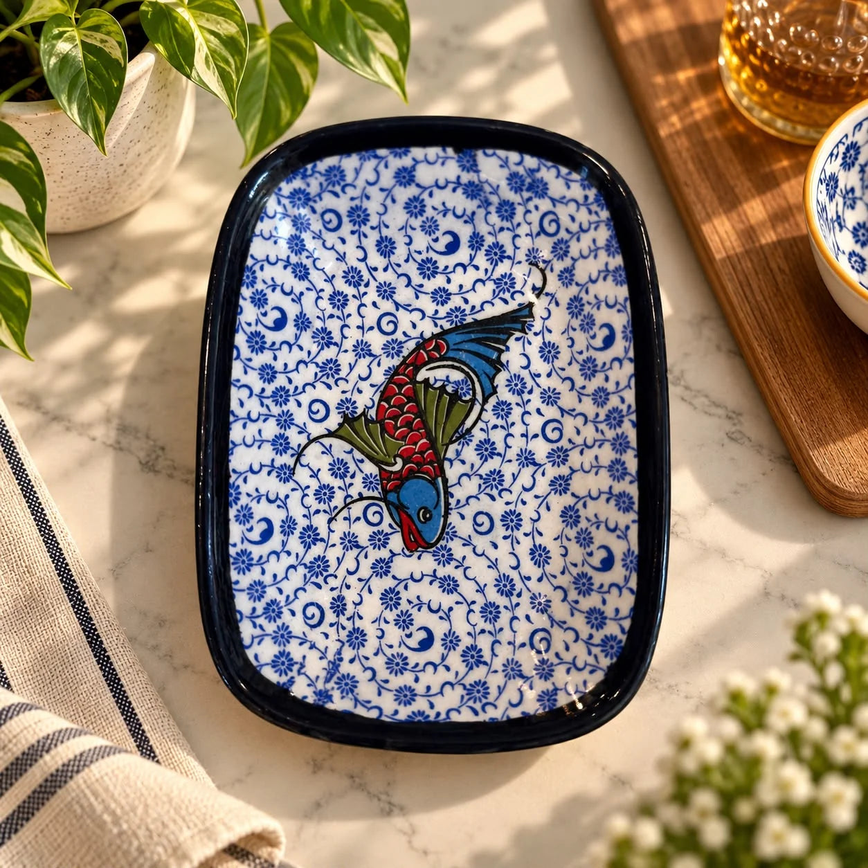 Plato rectangular de cerámica con borde negro, flores azules pequeñas y un pez estilizado rojo y azul